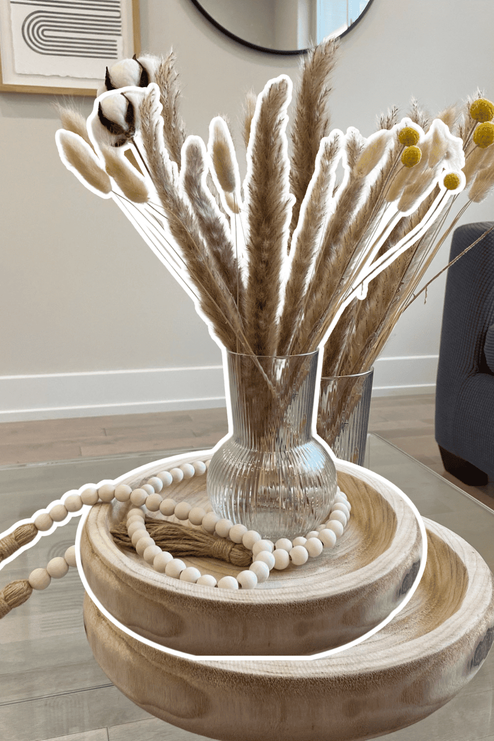 Elegant home decor featuring pampas grass in a glass vase with wooden accents and beaded accessories.